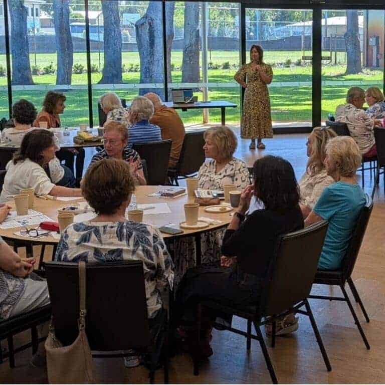 Community gathering with women seated around tables, listening to a speaker in a bright, modern room with large windows.