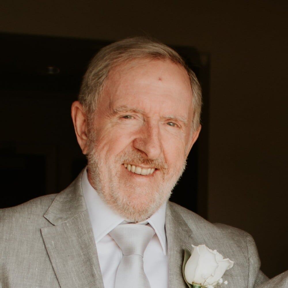 Smiling mature man in a light gray suit with a white rose boutonniere, indoor setting.