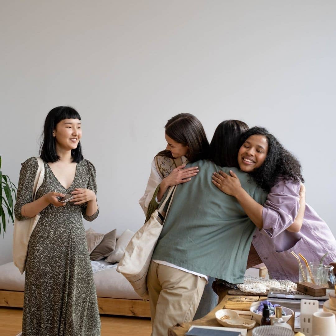 Group of diverse women hugging and smiling in a cozy indoor setting.